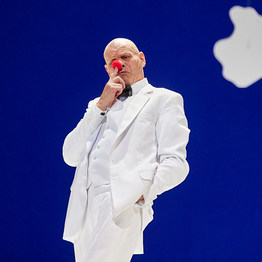 Szene aus der Oper »Die Nase«: Ein Mann im weißen Anzug steht vor einem blauen Hintergrund mit einer weißen Wolke und hält sich eine runde, rote Clownsnase an die Nase