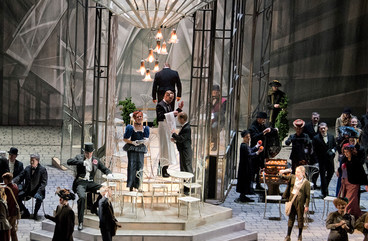 Szene aus "La Bohème": Mittig steht ein gläserner Kasten, drinnen und draußen stehen Café-Stühle. Einige wenige Sänger*innen befinden sich im Kasten, der Großteil steht außerhalb 
