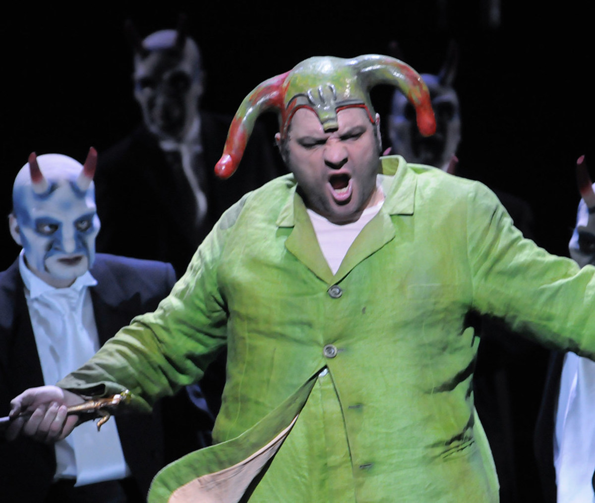Szene aus "Rigoletto": Sänger in grünfarbenem Kostüm steht vor weiteren Sängern