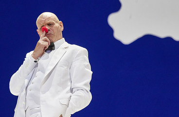 Szene aus der Oper »Die Nase«: Ein Mann im weißen Anzug steht vor einem blauen Hintergrund mit einer weißen Wolke und hält sich eine runde, rote Clownsnase an die Nase