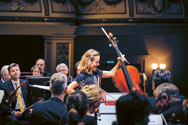 Die Cellistin Sol Gabetta steht mit erhobenem Cello zwischen den Musikerinnen und Musikern der Sächsischen Staatskapelle Dresden auf der Bühne der Semperoper.