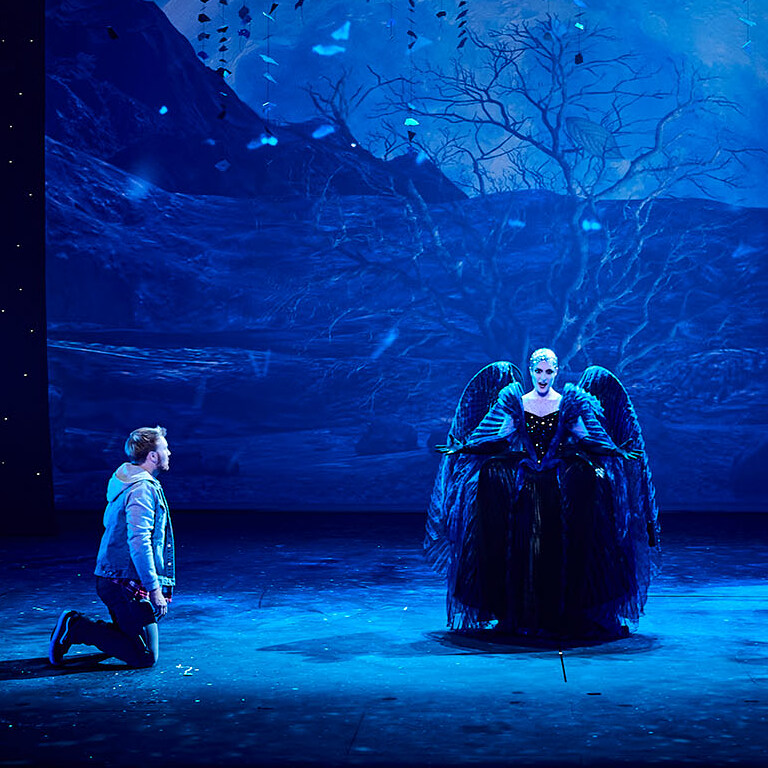 Header zauberfloete 2020 1680x836 Szene aus "Die Zauberflöte": Sängerin steht mittig auf der Bühne, ein Sänger kniet links neben ihr