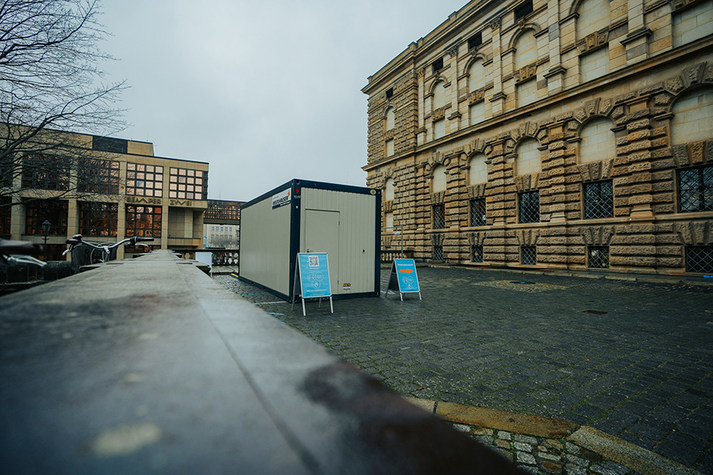 Ein grauer Container mit blau plakatierten Klappaufstellern davor steht auf einem Parkplatz neben der Semperoper