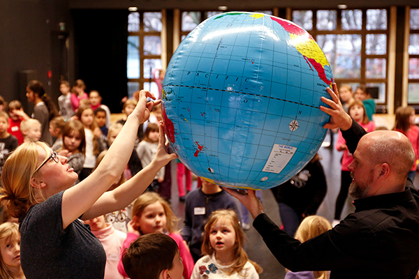 Zwei Musiktheater-Pädagog*innen zeigen einer staunenden Gruppe Kinder Orte auf einer großen aufblasbaren Weltkugel